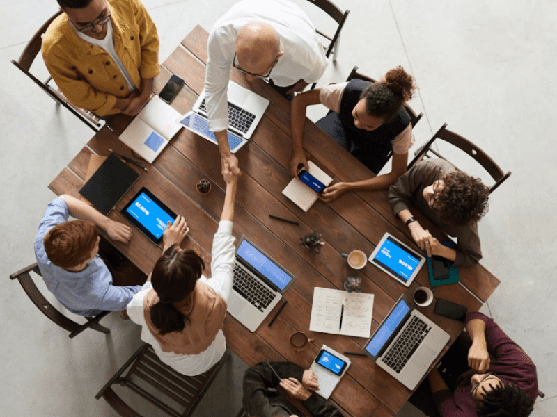 Trustack MSP Cyber Security, IT Services, IT Support. A group of eight people sit around a wooden table engaged in a Connectwise-driven meeting. Laptops, notebooks, and smartphones are visible on the table. Two individuals in the center are shaking hands. The setting appears to be a collaborative workspace or office.