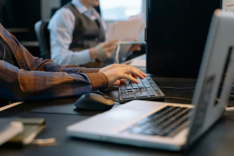 Trustack MSP Cyber Security, IT Services, IT Support. Close-up of a person typing on a keyboard at a desk with a laptop in the foreground, branded as a Dell Partner. Another individual is visible in the background, holding papers and sitting at a desk. The scene is set in an office environment.