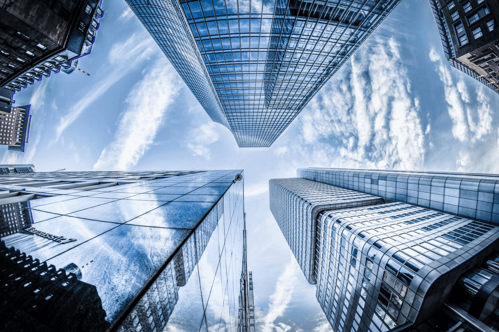 A fisheye lens captures the view from below of several towering skyscrapers reflecting the blue sky and clouds. The buildings converge towards the center, creating dramatic angles and showcasing modern architectural design, reminiscent of Trustack's innovative approach in EMEA Partner ventures.
