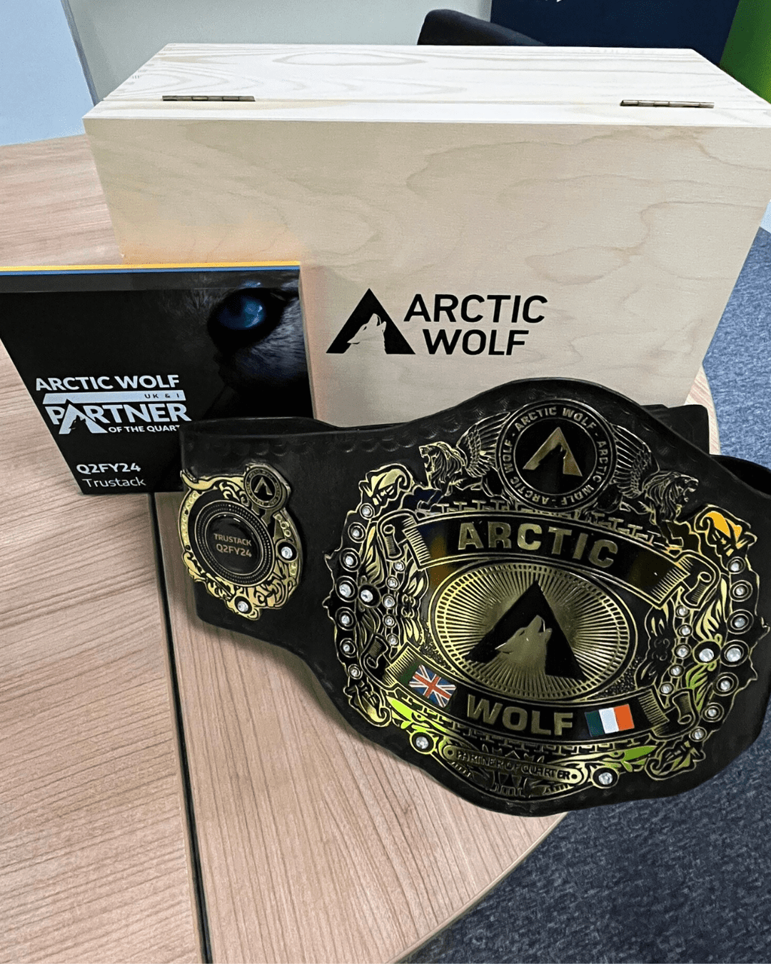 A championship-style belt and a box labeled "Arctic Wolf" are displayed on a wooden table. The belt features elaborate gold detailing and the Arctic Wolf logo. An upright booklet titled "Arctic Wolf EMEA Partner of the Quarter Q2 FY24" is beside the belt.
