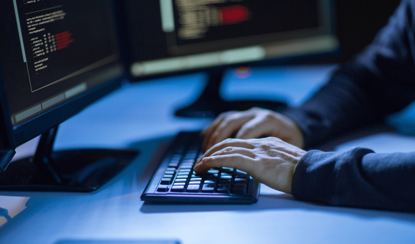 Trustack MSP Cyber Security, IT Services, IT Support. A person is typing on a computer keyboard with two monitors displaying code in the background. Their hands, visible and clad in a long-sleeved shirt, move like precision instruments. The environment resembles a recovery position for focus, dimly lit and illuminated by the glowing screens.