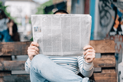 Trustack MSP Cyber Security, IT Services, IT Support. A person wearing a striped shirt and jeans reads Tech Industry Insights and News while sitting on a wooden bench outdoors. The person's face is hidden by the newspaper, blending into the urban setting with bikes, buildings, and other people in the background.