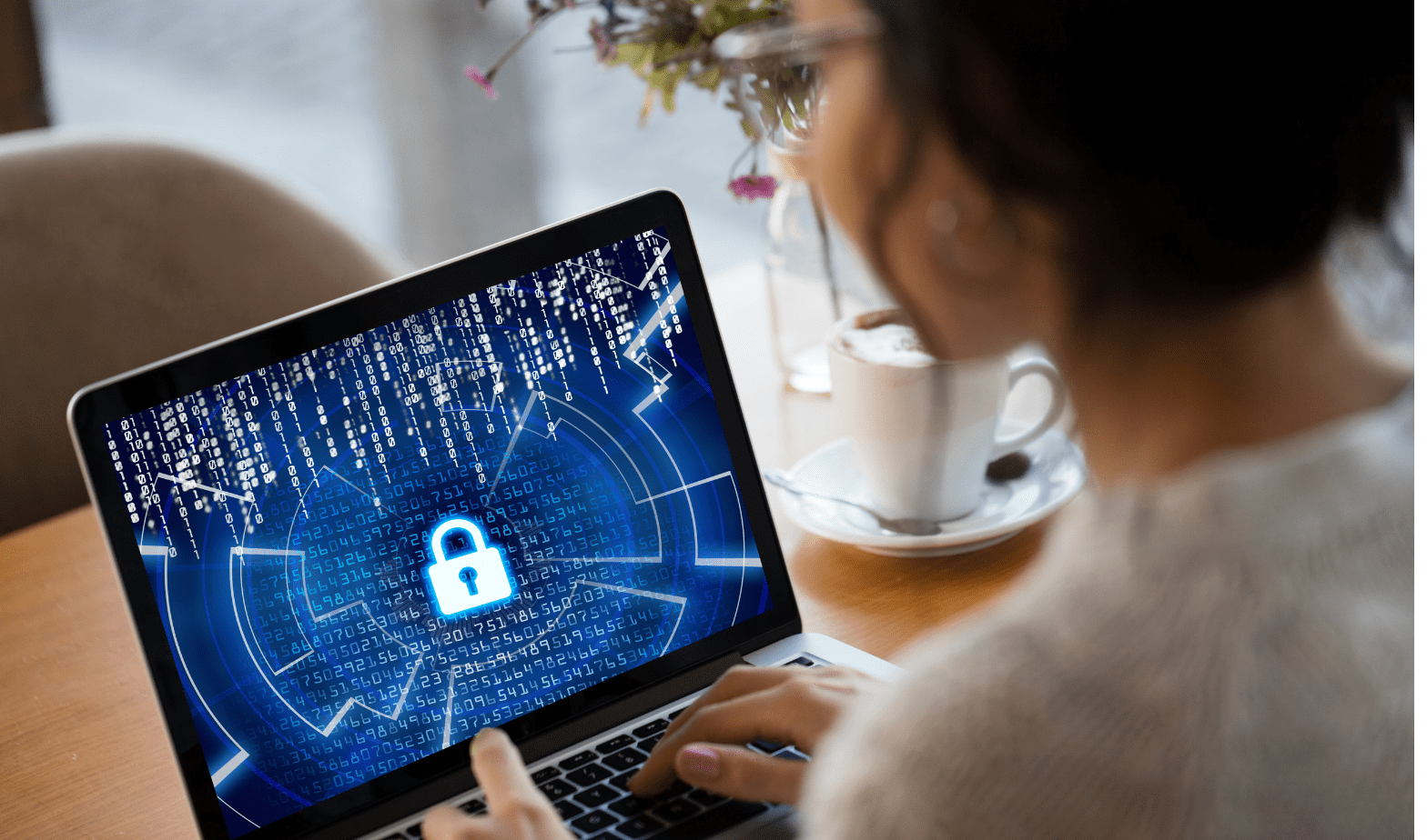 Trustack MSP Cyber Security, IT Services, IT Support. A person is sitting at a table using a laptop displaying a digital lock icon and binary code on the screen, symbolizing cybersecurity. The person is meticulously typing on the keyboard, aware that human error can compromise security. A cup of coffee and flower vase are in the background.