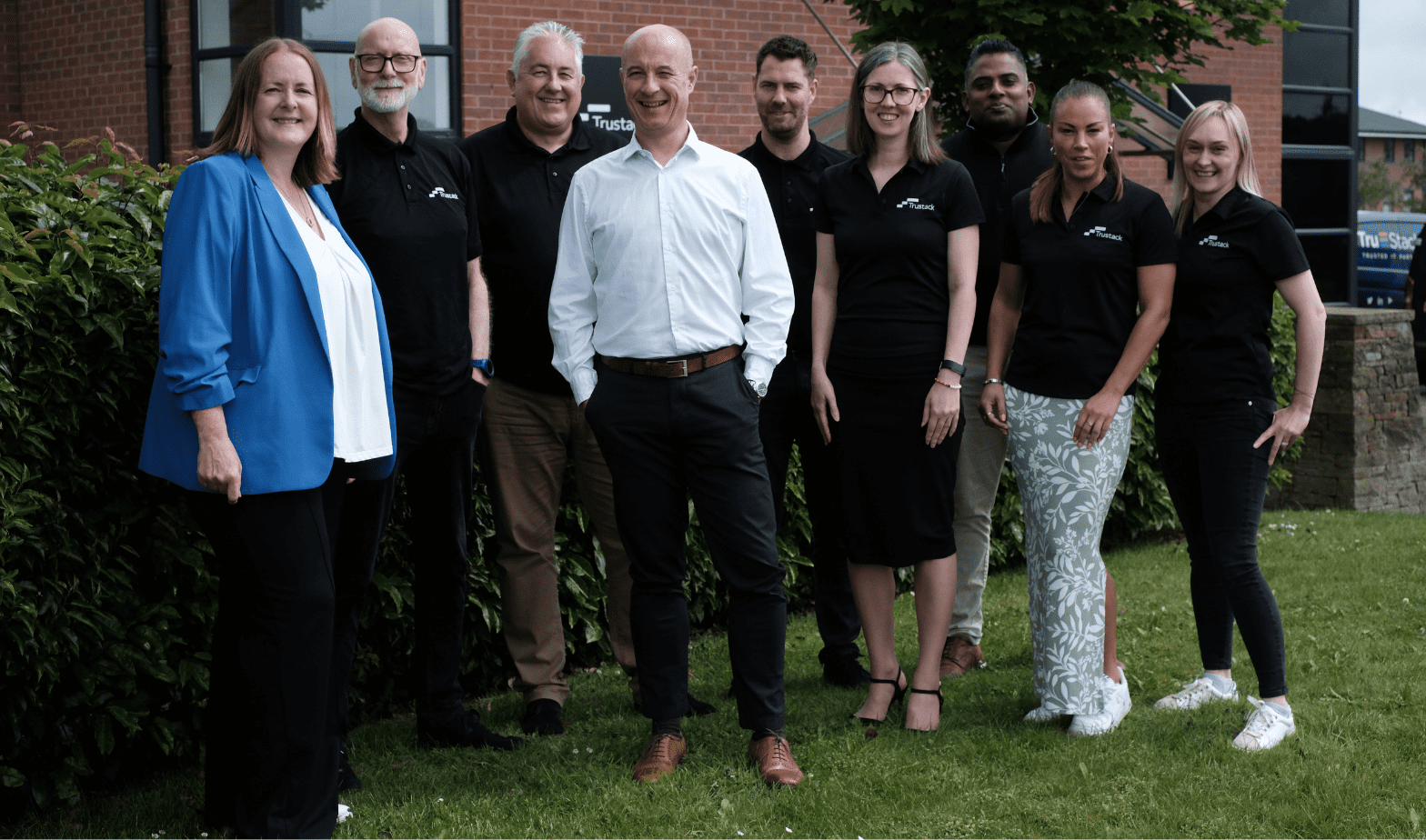 Trustack MSP Cyber Security, IT Services, IT Support. Nine individuals stand in a row, posing for a group photograph outdoors in front of a brick building. Four are wearing black shirts with a logo, one is in a white dress shirt, and another is in a blue blazer. The group stands on grass near some bushes. For more information about our team, please contact us.