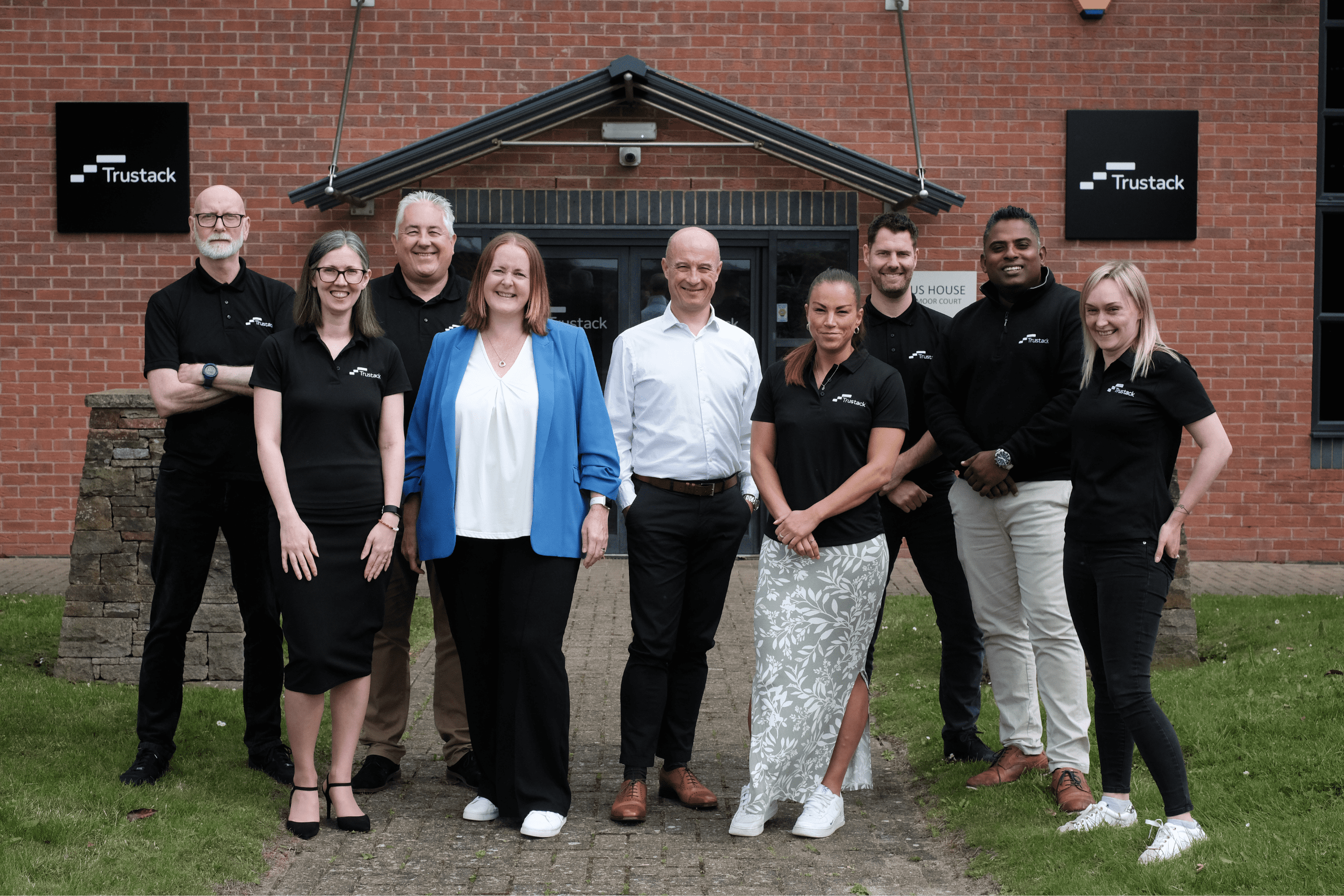 Trustack MSP Cyber Security, IT Services, IT Support. A group of ten people are standing in a line outside a brick building with two "Trustack MSP" signs. Some are dressed in black shirts, while others wear business casual. They are smiling and appear to be posing for a group photo, with foliage visible in the background.