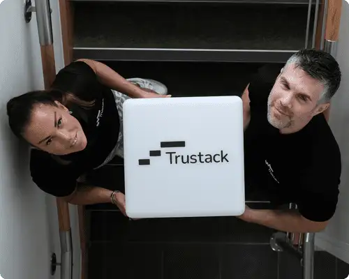 Trustack MSP Cyber Security, IT Services, IT Support. Two people on a staircase are holding a large white cube with the "Trustack" logo, representing the Trustack MSP. The photo is taken from above, with both individuals looking up towards the camera.