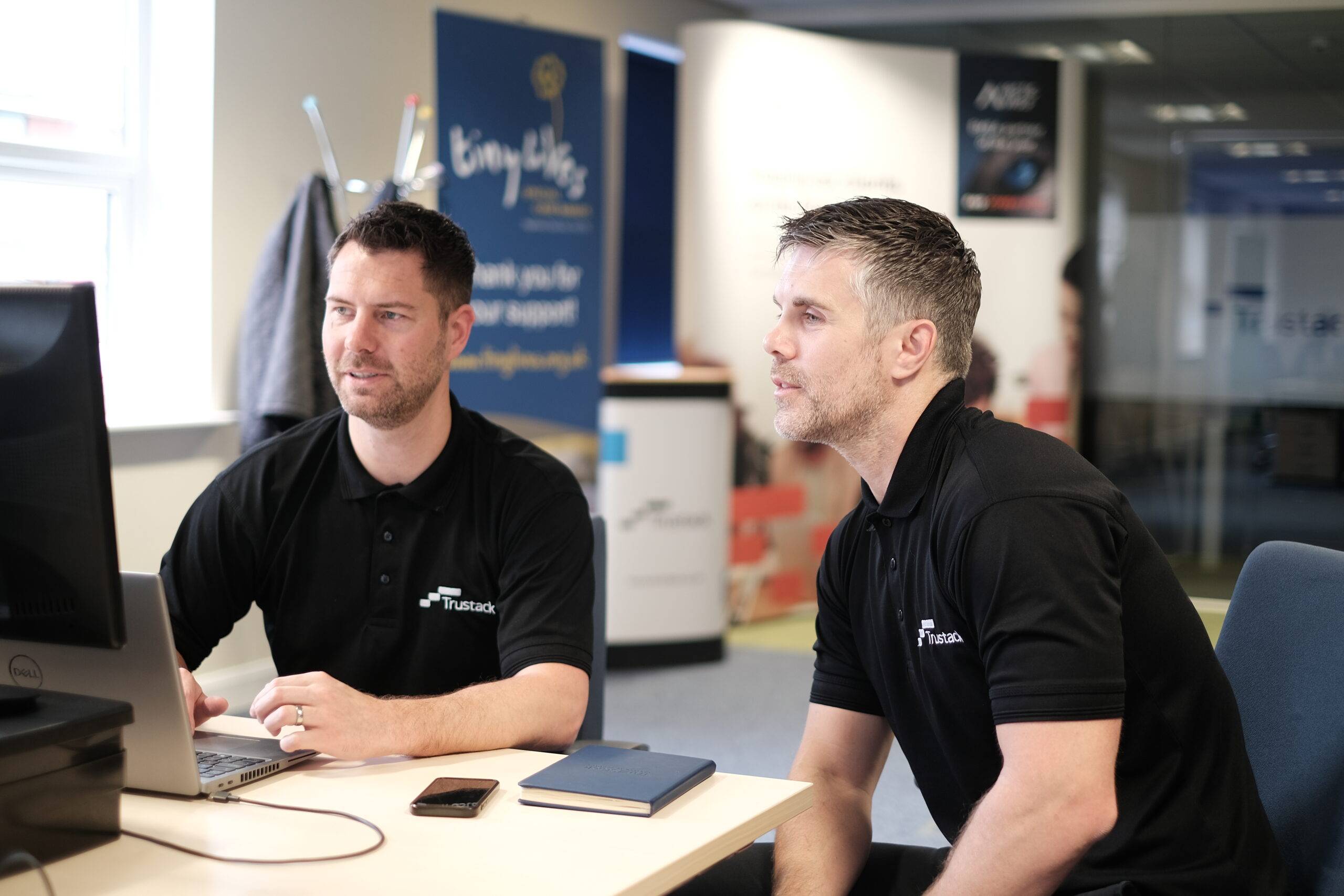 Trustack MSP Cyber Security, IT Services, IT Support. Two men in black polo shirts with "Trustack" logos sit at a desk, looking at a computer monitor. One man operates a laptop while the other sits beside him. A notebook and pen are on the desk.