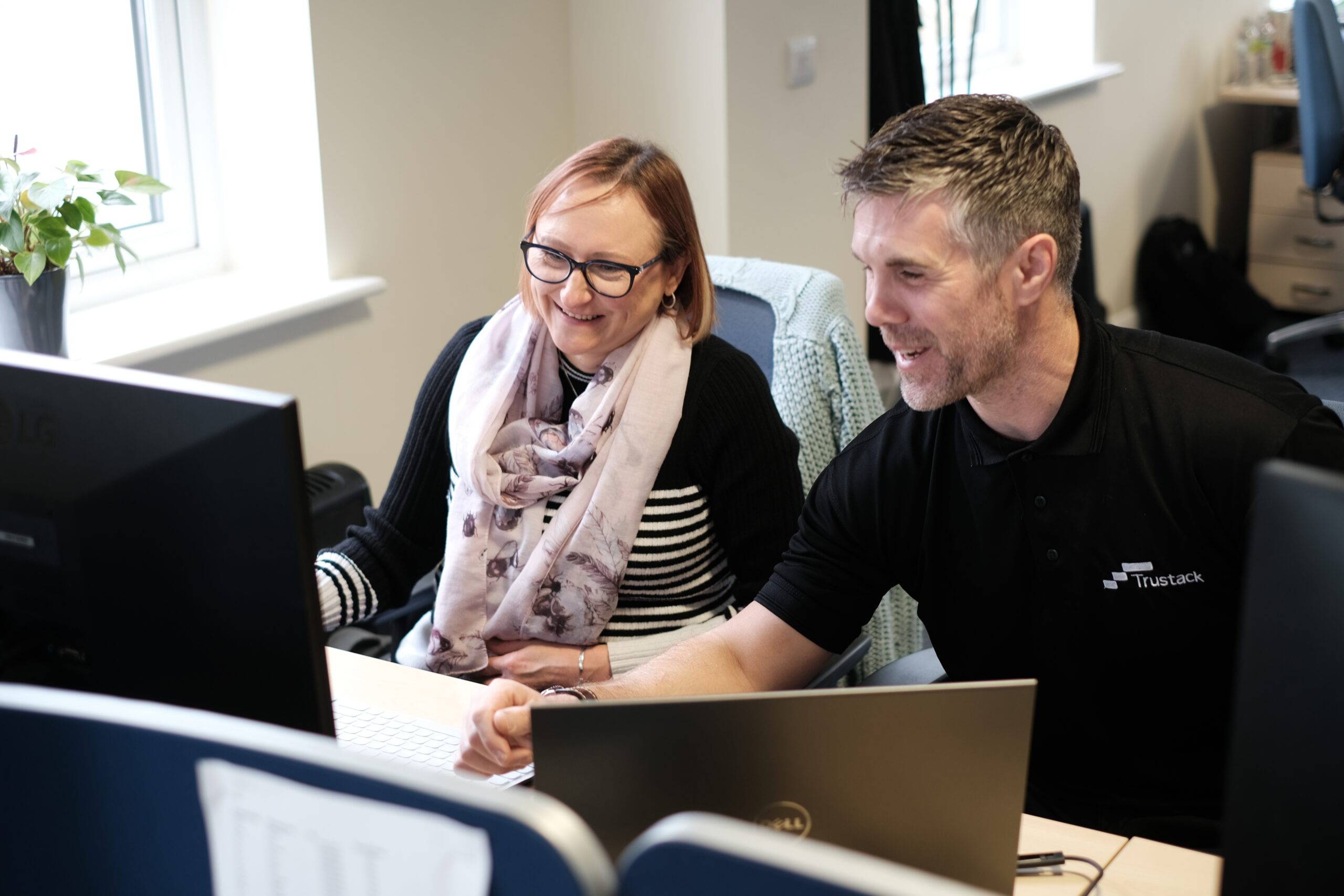 Trustack MSP Cyber Security, IT Services, IT Support. Two people sit side by side at a desk in an office, smiling as they review Managed Security solutions on a computer screen. The man points at the monitor while the woman, wearing glasses and a scarf, observes. Laptops and office furniture are visible.