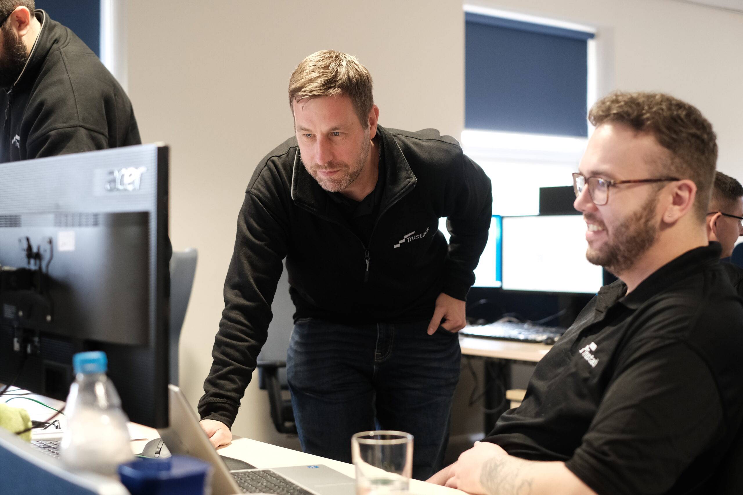 Trustack MSP Cyber Security, IT Services, IT Support. Two men in black shirts are working at a desk with computer screens. One man is sitting and smiling at his monitor, while the other is standing and leaning over to look at the screen. Office setting with multiple desks.