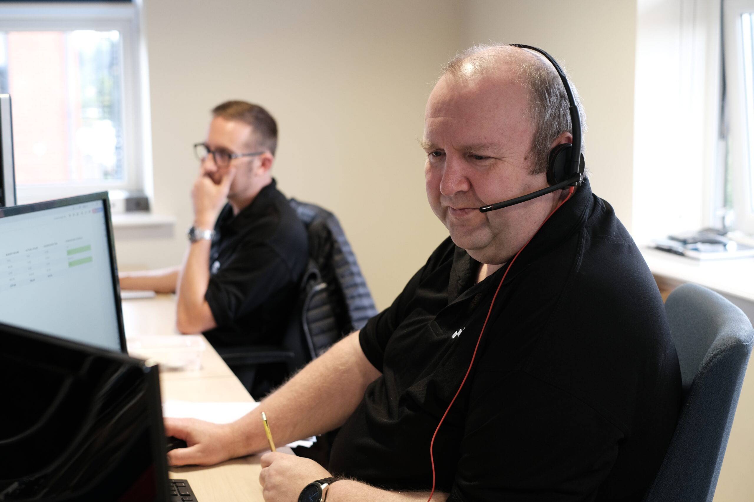 Trustack MSP Cyber Security, IT Services, IT Support. Two men sit at desks in an office, both wearing black shirts. The man in the foreground, focused on compliance tasks, wears a headset and looks at his computer screen, while the man in the background appears thoughtful, resting his hand on his chin.