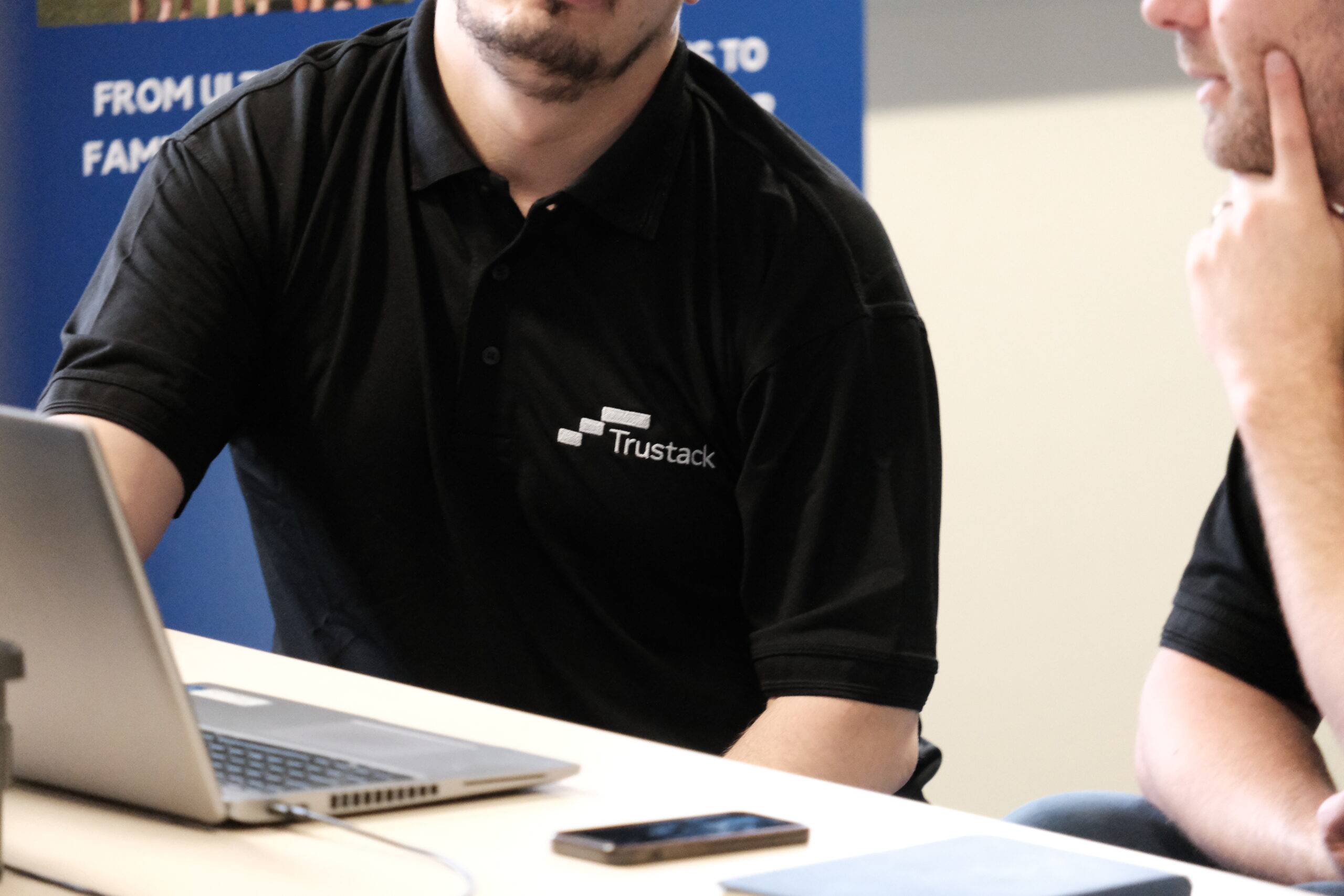 Trustack MSP Cyber Security, IT Services, IT Support. Two people wearing black polo shirts with a "Trustack" logo are seated at a table, focused on threat protection solutions displayed on a laptop and smartphone. One person has their hand on their chin, with a blue banner visible in the background.