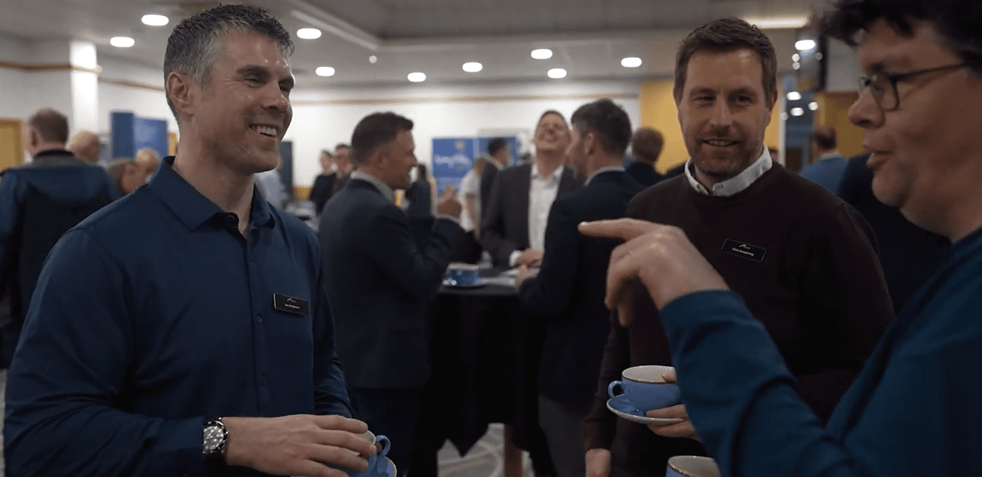Trustack MSP Cyber Security, IT Services, IT Support. Three men stand together holding coffee cups, engaged in conversation at a professional event. Other people are visible in the background, also talking and mingling in a well-lit room.