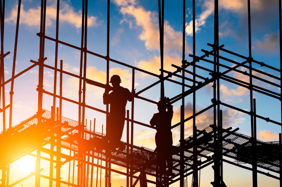 Trustack MSP Cyber Security, IT Services, IT Support. Two construction workers from the construction sector stand on scaffolding at a building site during sunset, with the sun low in the sky and a partly cloudy background.