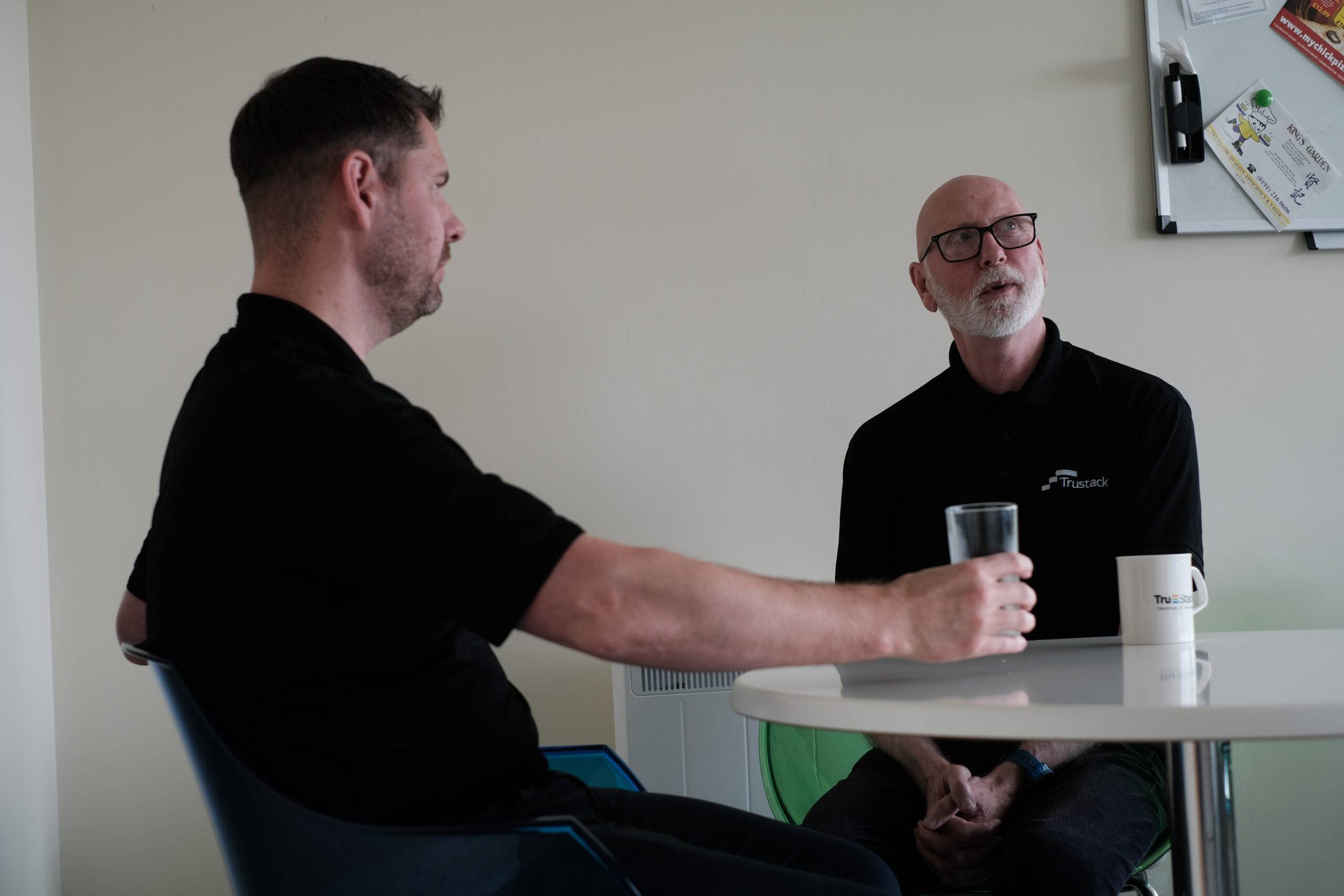 Trustack MSP Cyber Security, IT Services, IT Support. Two men sit at a small round table in an office break room. One holds a glass of water, the other has a mug. Both wear black shirts. A whiteboard with papers and a New Beyond Supported notice is visible on the wall behind them.