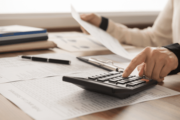 Trustack MSP Cyber Security, IT Services, IT Support. A person in the financial sector uses a calculator on a desk covered with financial documents, a clipboard, a pen, and folders. One hand holds a sheet of paper while the other presses buttons on the calculator.