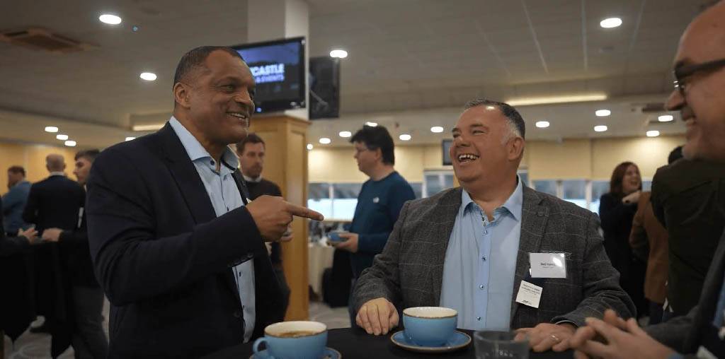 Trustack MSP Cyber Security, IT Services, IT Support. Two men in business attire stand at a table with coffee cups, smiling and talking at an indoor networking event. Other people are mingling in the background under bright ceiling lights.