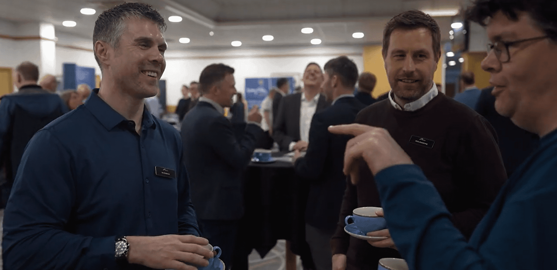 Trustack MSP Cyber Security, IT Services, IT Support. Three men stand talking and holding blue coffee cups at a networking event, with other attendees conversing in the background of a well-lit indoor venue.