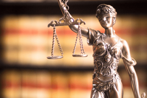 Trustack MSP Cyber Security, IT Services, IT Support. A close-up of a metallic statue of Lady Justice holding balanced scales, blindfolded, symbolizes fairness in the Legal Sector, with blurred shelves of books in the background.