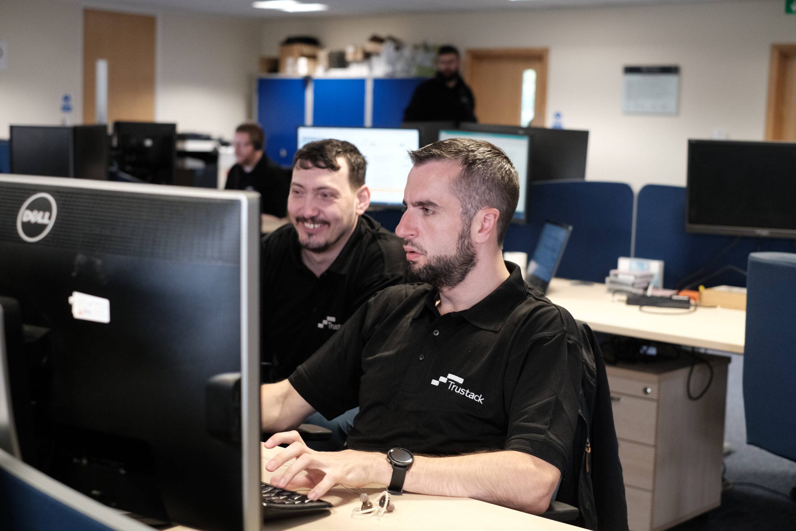 Trustack MSP Cyber Security, IT Services, IT Support. Two men wearing black polo shirts with the Trustack logo work at desktop computers in an office with desks, blue partitions, and other colleagues in the background.