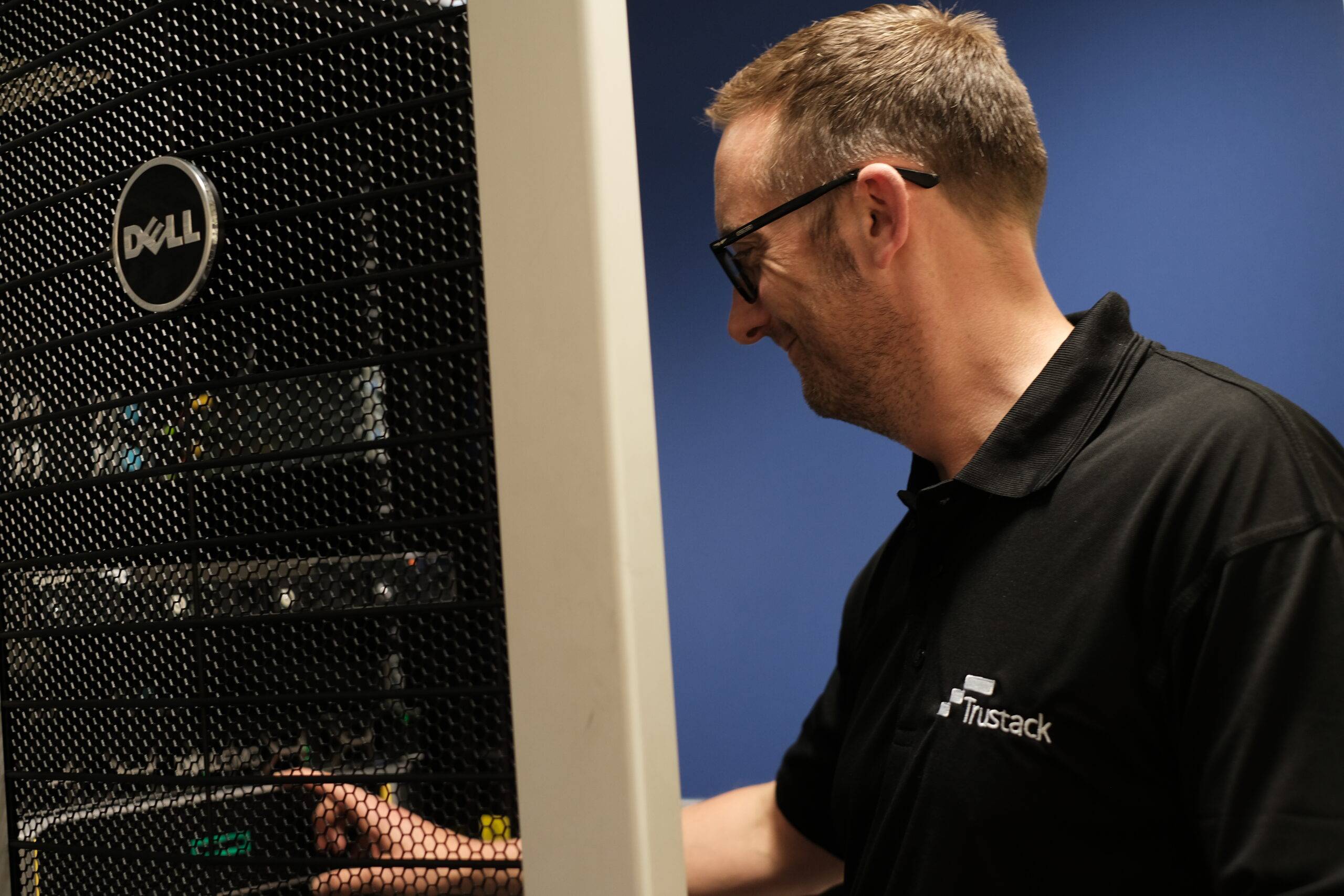 Trustack MSP Cyber Security, IT Services, IT Support. A man wearing glasses and a black "Trustack" polo shirt works on Networking Hardware, configuring a Dell server rack in a room with blue walls.