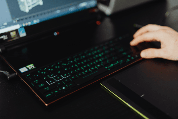 Trustack MSP Cyber Security, IT Services, IT Support. A person’s hand rests on a black keyboard with green backlighting, beside a graphics tablet and a laptop displaying a computer-aided design programme—illustrating seamless endpoint support in a creative workspace.