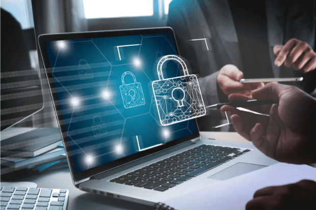 Trustack MSP Cyber Security, IT Services, IT Support. A laptop displays a digital padlock icon on the screen, symbolising Next-Gen Antivirus cyber security. In the background, two people are discussing something, with one holding a pen and the other a mobile phone.
