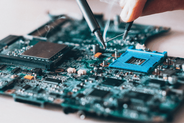 Trustack MSP Cyber Security, IT Services, IT Support. A close-up of a person using tools to solder components onto a computer motherboard, with visible smoke rising from the process, highlights the importance of precise Infrastructure Hardware work in building reliable digital systems.