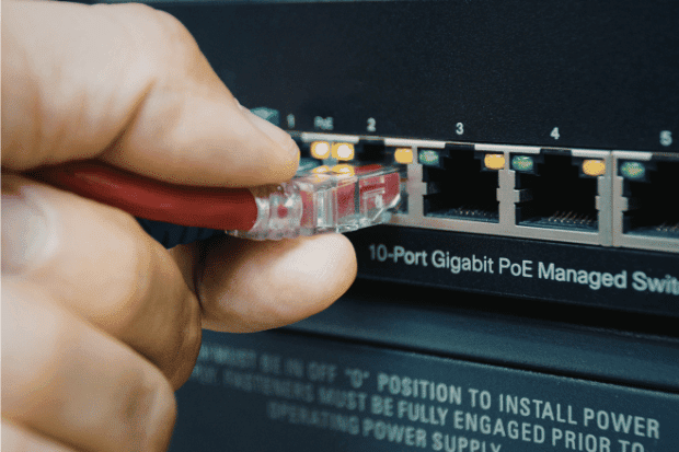 Trustack MSP Cyber Security, IT Services, IT Support. A close-up of a hand plugging a red Ethernet cable into a port on a 10-port Gigabit PoE managed network switch, with indicator lights visible above the ports, highlighting robust Switch Support.