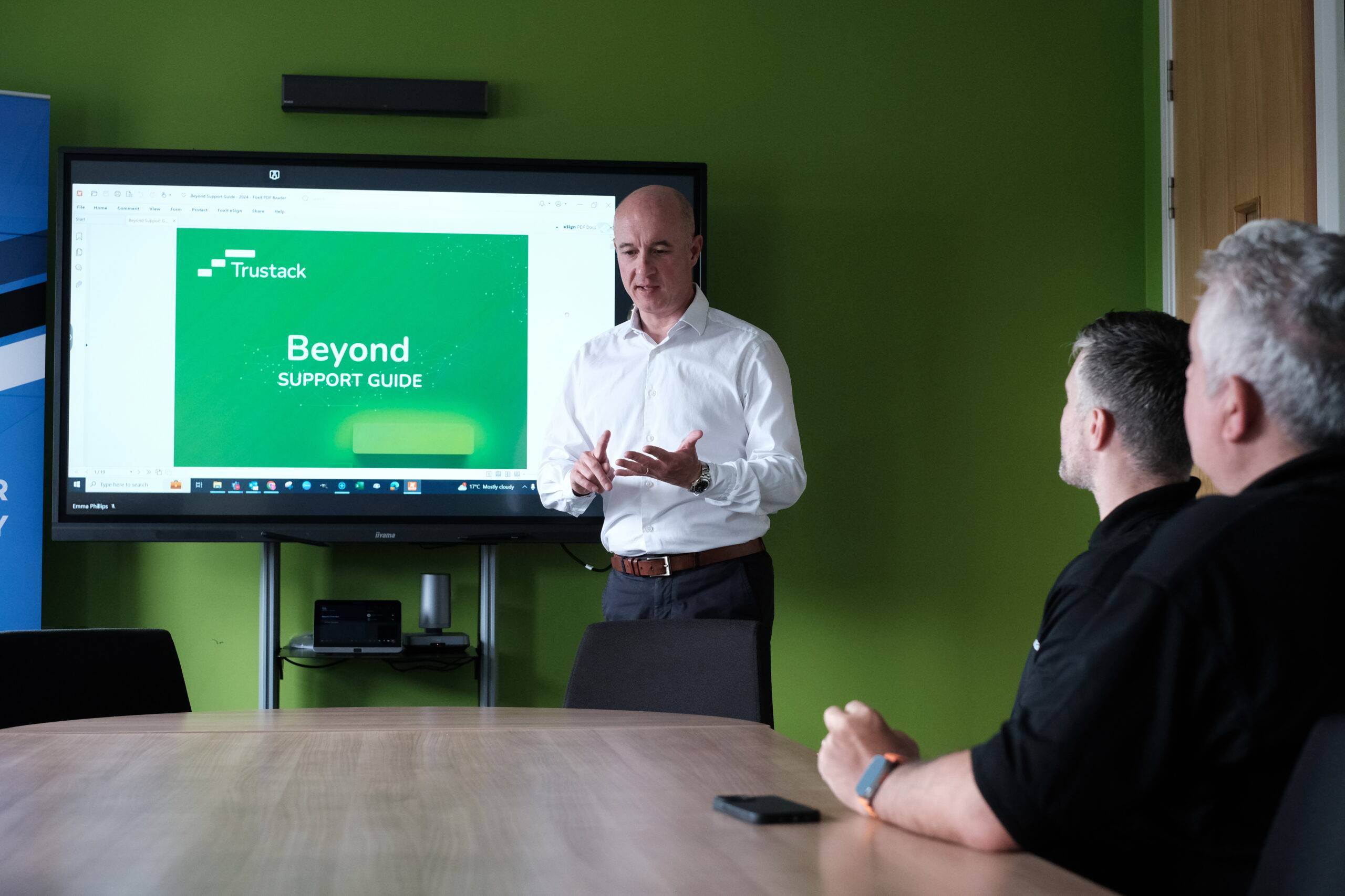 Trustack MSP Cyber Security, IT Services, IT Support. A man in a white shirt stands and gestures while presenting a slide titled “Trustack Beyond Support Guide” to two seated men in a conference room with green walls.