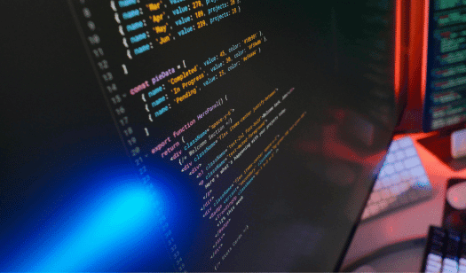 Trustack MSP Cyber Security, IT Services, IT Support. A computer monitor displays lines of colourful JavaScript code on a dark background. A keyboard and a blurred second screen complete the scene, evoking a development workspace—perfect for an Incident Commander Workshop setting.