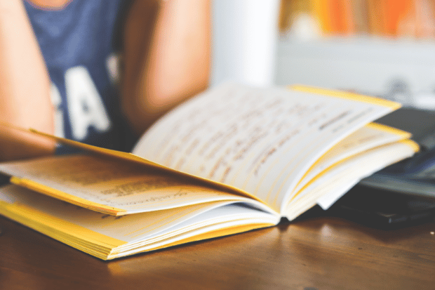 Trustack MSP Cyber Security, IT Services, IT Support. An open notebook with handwritten notes sits on a wooden table. A person with folded arms is partially visible in the background, wearing a sleeveless top. The scene suggests studying or working.