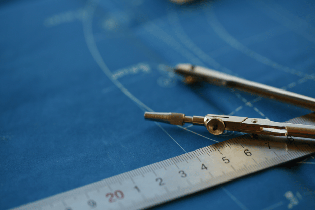 Trustack MSP Cyber Security, IT Services, IT Support. A close-up of a metal compass and a ruler placed on a blue architectural plan with white lines and measurements.