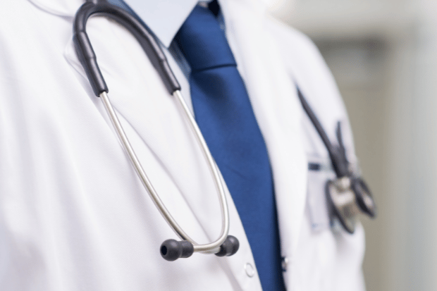 Trustack MSP Cyber Security, IT Services, IT Support. A close-up of a person wearing a white lab coat, blue dress shirt, and blue tie with a stethoscope draped around their neck, suggesting a medical professional.