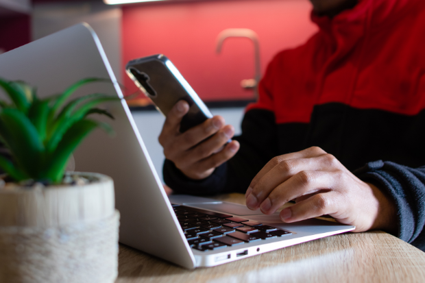 Trustack MSP Cyber Security, IT Services, IT Support. A person in a red and black jacket uses a mobile phone for authentication while typing on a laptop with the other hand, seated at a wooden table with a small potted plant nearby.