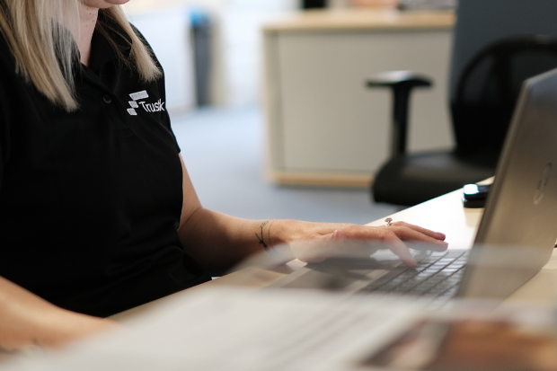 Trustack MSP Cyber Security, IT Services, IT Support. A person with fair hair wearing a black polo shirt sits at a desk typing on a laptop. The shirt has a logo and the word "Tusk." An office chair and desk are visible in the background, suggesting work related to access control.
