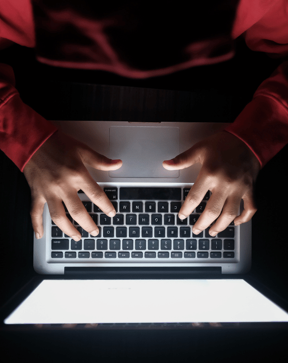Trustack MSP Cyber Security, IT Services, IT Support. A person in a red long-sleeve shirt types on a laptop keyboard, illustrating the importance of DLP in business security. The image, taken from above, shows the screen’s white light illuminating the hands and keys.