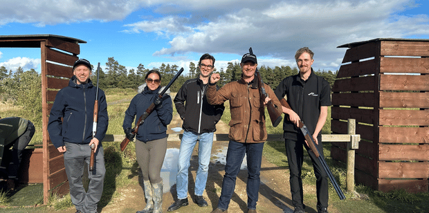 Trustack MSP Cyber Security, IT Services, IT Support. Five people stand outdoors at a clay shooting range, each holding a shotgun. Dressed casually, they are surrounded by wooden dividers and grassy terrain, with a partly cloudy sky completing the BlackPoint Clay Pigeon Shoot scene.