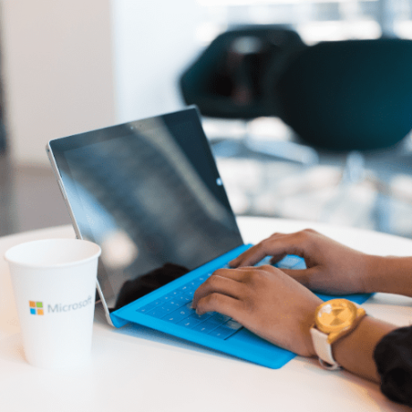 Trustack MSP Cyber Security, IT Services, IT Support. A person is typing on a blue keyboard attached to a tablet device, likely using an lp-template. The setting appears to be an office with modern seating in the background. A white paper cup with the Microsoft logo is placed on the table beside the device.