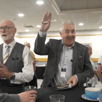 Trustack MSP Cyber Security, IT Services, IT Support. Four men stand around a round table at a professional event. One man smiles and raises his hand, holding a pen. The table has empty glasses and a cup. Office lighting and other attendees are visible in the background.